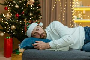 uma homem é cansado para Natal, vestindo uma Novo anos chapéu, dormindo perto a Natal árvore, sentado em a sofá às casa dentro a vivo quarto depois de a comemorar Natal. foto