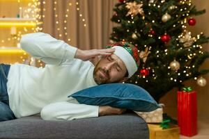 uma homem é cansado para Natal, dentro uma Novo anos chapéu, ele é dormindo de a Natal árvore em a sofá às casa dentro a vivo quarto depois de a comemorar Natal, ele tem uma mau dor de cabeça a partir de uma ressaca. foto