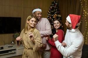 quatro diverso amigos às Novo anos festa a comemorar e dançando, casa convidados tendo Diversão às Natal, dentro Novo anos festivo roupas e santa chapéus dentro a vivo sala. foto