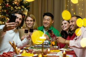uma companhia do interracial pessoas a comemorar a Novo ano junto, sentado às a festivo mesa. a homem é segurando a telefone, todos é falando em uma vídeo ligar com amigos, saudações, sorridente. foto