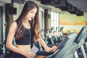 mulher asiática usando telefone inteligente quando treino ou treinamento de força no ginásio de fitness na esteira. relaxe e conceito de tecnologia. exercício esportivo e tema de cuidados de saúde. humor feliz e confortável foto