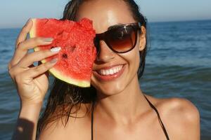 verão período de férias - jovem menina comendo fresco Melancia em arenoso de praia foto