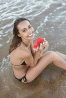 verão período de férias - jovem menina comendo fresco Melancia em arenoso de praia foto