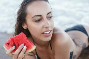 verão período de férias - jovem menina comendo fresco Melancia em arenoso de praia foto