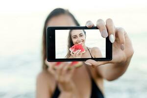 verão período de férias - jovem menina comendo fresco Melancia em arenoso de praia foto