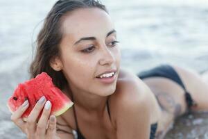 verão período de férias - jovem menina comendo fresco Melancia em arenoso de praia foto