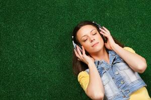 lindo jovem mulher com fones de ouvido goza dentro música enquanto deitado baixa em Relva foto