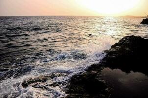 a Sol é configuração sobre a oceano Como ondas batida contra a pedras foto