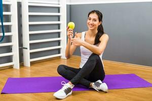 lindo ginástica mulher comendo maçã e mostrando polegar acima às a Academia foto