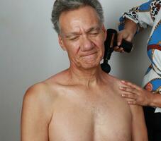 Mais velho maduro homem tendo uma massagem em dele superior costas e pescoço para dor alívio com uma percussão massagem arma de fogo foto