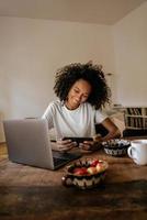 jovem negra usando telefone celular e laptop enquanto toma o café da manhã foto