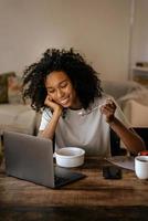 jovem negra com fones de ouvido usando laptop e tomando café da manhã