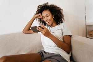 jovem negra com fones de ouvido usando o celular enquanto está descansando no sofá