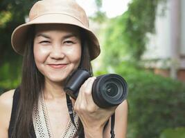 mulher asiática, usando chapéu e top preto sem mangas, de pé no jardim e segurando a câmera dslr, sorrindo alegremente. foto