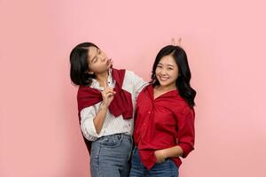 ásia fêmea amigos rindo e posando em Rosa fundo uma lindo sorridente mulher vestindo uma vermelho camisa é feliz. uma alegre menina vestindo uma branco camisa dá Coelho orelhas para amigo foto