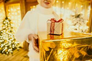 mulher dentro branco casaco segurando embrulhado em ouro presente caixa com vermelho fita dentro frente do Natal árvore, implicando feriado estação presente intercâmbio. foto