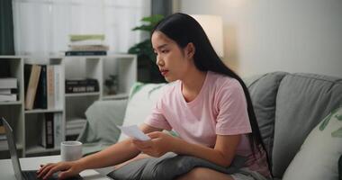 retrato do jovem ásia mulher olhando às computador portátil tela segurando fatura recibo dentro mão planejamento família despesas comparando papel e conectados documento, casa finança foto