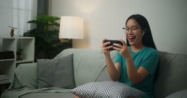 retrato do jovem ásia mulher vestindo óculos e fones de ouvido goza jogando conectados esport jogos em Smartphone sentado em sofá dentro a vivo quarto às casa, jogador estilo de vida conceito. foto