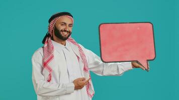 muçulmano adulto detém discurso bolha em Câmera, criando propaganda para Novo rede comercial. jovem pessoa trabalhando em marketing indústria com esvaziar cartão ícone, isolado Painel publicitário placa. foto