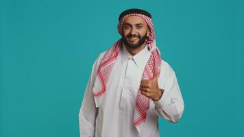 meio Oriental homem faz polegares acima em Câmera, mostrando acordo e positividade enquanto ele desgasta tradicional islâmico roupas. jovem sorridente cara dentro nacional árabe vestuário, gostar símbolo. foto