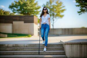 ai gerado uma cego ásia jovem mulher vestindo oculos de sol anda em baixa alguns passos com uma caminhando bastão dentro dela mão e uma branco camisa em topo do dela foto