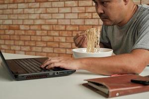 trabalhador tailandês do sexo masculino ocupado trabalhando com laptop, use os pauzinhos para comer macarrão instantâneo apressadamente durante o intervalo do almoço do escritório, porque é rápido, saboroso e barato. ao longo do tempo fast food asiático, estilo de vida pouco saudável. foto