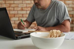 trabalhador tailandês do sexo masculino ocupado trabalhando com laptop, use os pauzinhos para comer macarrão instantâneo apressadamente durante o intervalo do almoço do escritório, porque é rápido, saboroso e barato. ao longo do tempo fast food asiático, estilo de vida pouco saudável. foto