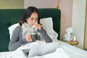 retrato do ásia menina espirros e limpando escorrendo nariz, sentindo-me doente, ficando dentro cama com gripe, olhando às computador portátil foto