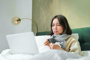 retrato do jovem ásia mulher deitado doente dentro cama, ficando às casa com frio ou gripe, assistindo vídeos, usando computador portátil dentro dela quarto foto
