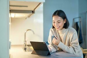 retrato do à moda jovem ásia mulher assistindo videos em tábua, sentado dentro cozinha e bebendo café foto