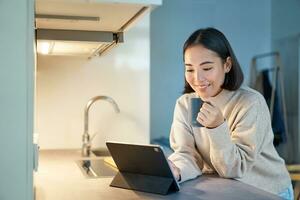 retrato do à moda jovem ásia mulher assistindo videos em tábua, sentado dentro cozinha e bebendo café foto