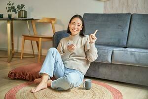 sorridente ásia menina senta em chão dentro à moda vivo sala, apontando dedo às anúncio, mostrando promo bandeira, segurando Móvel telefone dentro mão foto