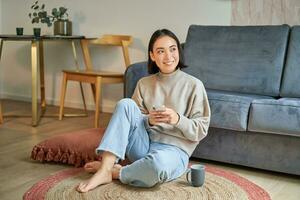 imagem do à moda jovem mulher dentro moderno casa, usando Móvel telefone, sentado em chão e segurando Smartphone, bebendo a partir de copo foto