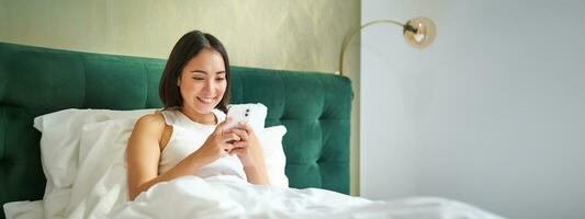 lindo sorridente ásia mulher dentro cama, lendo mensagem em Smartphone e rindo, desfrutando preguiçoso dia dentro quarto, usando Móvel telefone foto