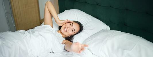 romântico ásia menina acorda acima e alongar, sorridente Felizmente, alcançando dela mão, alongamento braço para você, deitado dentro cama dentro branco folhas foto