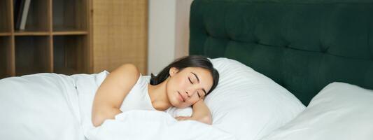 lindo ásia menina dorme dentro dela cama com branco folhas, caloroso inverno edredon, sorridente dentro dela dormir, sonhando, deitado dentro quarto dentro manhã foto