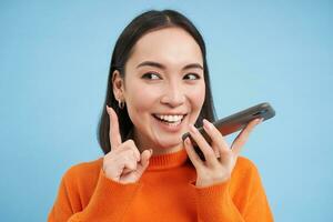 retrato do coreano mulher registros voz mensagem em Smartphone aplicativo, fala em palestrante, traduz dela discurso em Móvel aplicativo, azul fundo foto