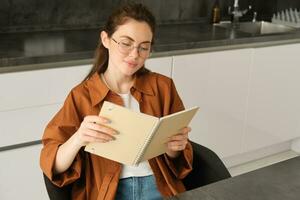 fechar acima retrato do jovem mulher, aluna dentro cozinha, segurando caderno, revisando para exame às lar, estudo, lendo dela planejador foto