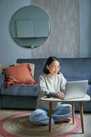 vertical tiro do coreano trabalhando mulher, sentado em chão às casa com computador portátil, estudo, usando computador em controlo remoto, e-learning conceito foto