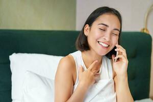 fechar acima retrato do fofa ásia menina dentro cama, falando em Móvel telefone com feliz sorridente face. mulher acordando acima e fazer uma Telefone ligar foto