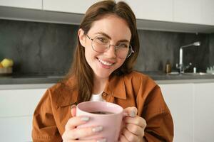 fechar acima retrato do lindo mulher dentro copos, bebidas chá, aquece dela mãos com quente xícara, sorridente e olhando feliz às Câmera, sentado dentro cozinha foto