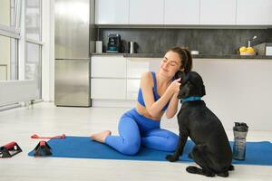 retrato do lindo morena ginástica garota, tocam com dela cachorro durante exercite-se Treinamento sessão, senta às casa em borracha ioga esteira, faz esporte exercícios dentro vivo quarto foto