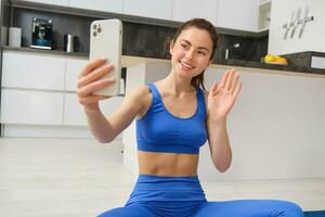 sorridente lindo menina registros vídeo enquanto fazendo Esportes às lar, parece às Smartphone, leva selfie em Móvel telefone, exercite-se dentro de casa dentro azul perneiras e sportsbra foto