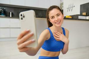 sorridente lindo menina registros vídeo enquanto fazendo Esportes às lar, parece às Smartphone, leva selfie em Móvel telefone, exercite-se dentro de casa dentro azul perneiras e sportsbra foto