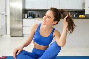 retrato do jovem, saudável e em forma morena mulher, senta em borracha esteira, faz ioga, exercite-se Treinamento às lar, desgasta azul ginástica roupa de esporte e sorrisos foto