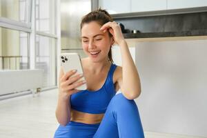 imagem do jovem em forma mulher vestindo sutiã esportivo e azul perneiras, usando Smartphone aplicativo, faz exercite-se a partir de lar, senta em chão e segue conectados Treinamento vídeo foto