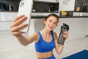 retrato do desportista leva selfie com água garrafa dentro vivo sala, detém Smartphone e poses para foto enquanto fazendo ginástica exercite-se