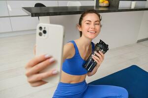 retrato do mulher fazendo Esportes, leva selfie em Smartphone, ginástica instrutor registros dela exercícios, permanece hidratado, bebidas água durante Treinamento sessão foto