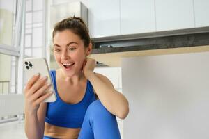 fechar acima tiro do jovem feliz mulher, fazendo Esportes, exercite-se às lar, verificação dela telefone, olhando espantado às Smartphone tela foto