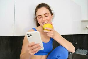 retrato do desportista, menina comendo e maçã e olhando às dela social meios de comunicação, Smartphone tela, tendo uma lanche dentro cozinha, vestindo ginástica roupa ativa foto